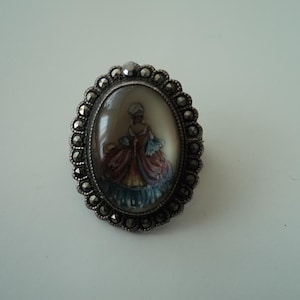 Op de afbeelding: Een ovale zilveren broche met een cameo van een vrouw in een kleurrijke jurk. De broche is omgeven door een rand van kleine, zwarte stenen.