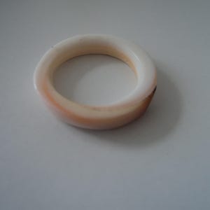 Puede incluir: Un anillo circular liso que presenta un degradado de colores que transita desde un blanco crema en la parte superior hasta un tono melocotón suave y marrón claro a lo largo del borde inferior. La banda tiene un perfil grueso y redondeado.