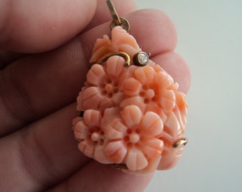 May include: A carved coral pendant in the shape of a cluster of flowers. The pendant is a light peach color with gold accents and a small diamond. The floral design is detailed and intricate.