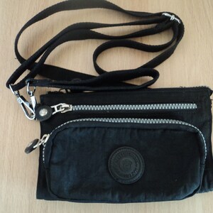 Puede incluir: Un bolso bandolera de nylon negro con cierre de cremallera y un broche de metal plateado. El bolso tiene un pequeño bolsillo en la parte delantera con un logotipo circular.