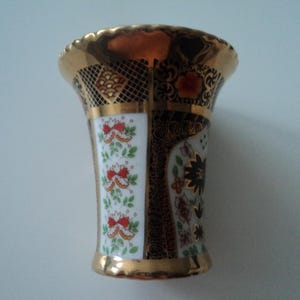 Puede incluir: Un pequeño jarrón decorativo con borde y base dorados. El jarrón presenta un fondo blanco con diseños florales rojos y un borde negro y dorado. El borde superior tiene un ribete dorado.