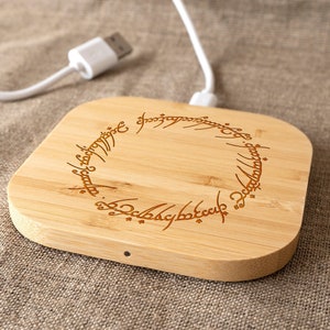 Può includere: Un caricabatterie wireless in legno con un design circolare che presenta l'iscrizione "Un anello per domarli tutti" in scrittura elfica.