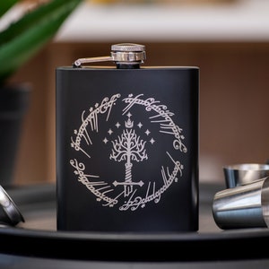 May include: Black stainless steel flask with a silver engraving of the One Ring from The Lord of the Rings. The engraving features the inscription "One Ring to rule them all, One Ring to find them, One Ring to bring them all and in the darkness bind them."