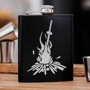 May include: Black metal flask with a silver design of a sword and flames. The design is reminiscent of a bonfire.