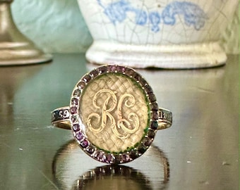 May include: A gold ring with a circular face displaying the intertwined letters "RG" in a script font. The ring's face is encircled by small purple gemstones. The ring is set against a dark, reflective surface.
