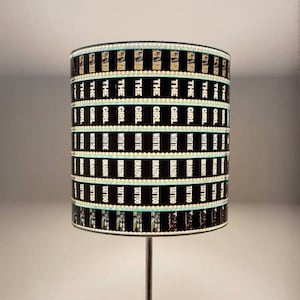 Può includere: Lampada da tavolo argentata con paralume cilindrico nero. Il paralume presenta la scritta bianca "THE GIRL WITH THE DRAGON TATTOO". La lampada è posizionata su un tavolo di legno.