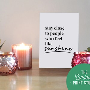 May include: A white card with the text "stay close to people who feel like sunshine" in black lettering. The card is sitting on a wooden table with two candles and two small plants.