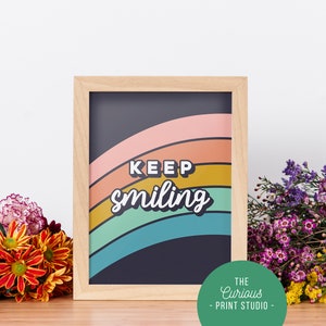 May include: A framed print with a rainbow design and the text "KEEP smiling" in a retro font. The frame is light wood and the background is dark blue. The print is from The Curious Print Studio.