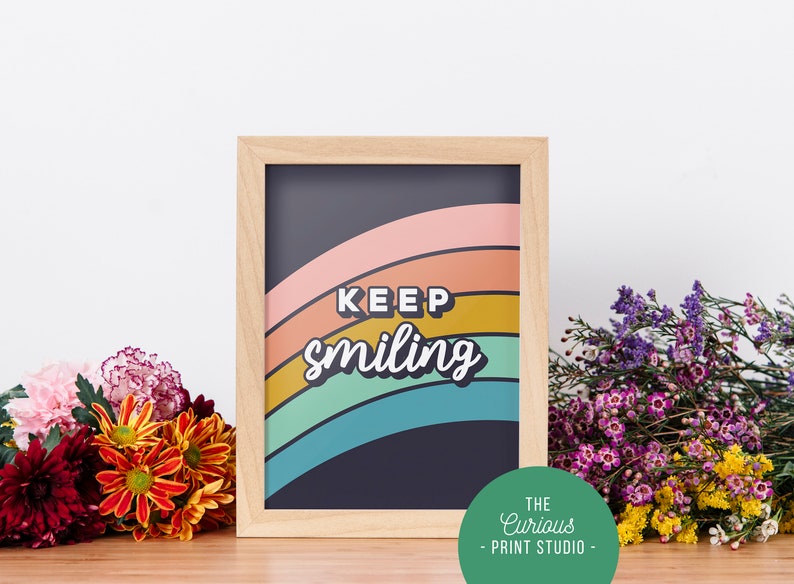 May include: A framed print with a rainbow design and the text "KEEP smiling" in a retro font. The frame is light wood and the background is dark blue. The print is from The Curious Print Studio.