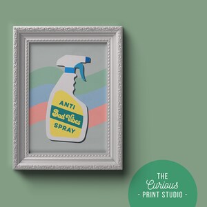 May include: A white framed print featuring a colourful illustration of a spray bottle labelled "Anti Bad Vibes Spray". The background is a pastel blue, pink and green striped pattern.