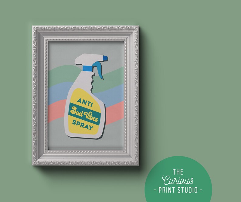 May include: A white framed print featuring a colourful illustration of a spray bottle labelled "Anti Bad Vibes Spray". The background is a pastel blue, pink and green striped pattern.