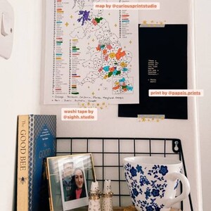 May include: A white wall with a colorful map of the UK sales map, a black framed print with white text, a wooden shelf with a gold framed photo, two ceramic salt and pepper shakers, a blue and white floral teacup, and a book titled "The Good Bee".