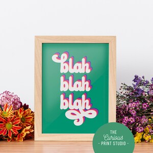 Puede incluir: Una impresi&oacute;n enmarcada con la palabra "blah" repetida tres veces en una fuente en capas. El texto es blanco con sombras rosas y moradas, sobre un fondo verde azulado. El marco es de madera clara. Las flores rodean la impresi&oacute;n.