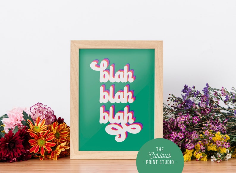 Puede incluir: Una impresi&oacute;n enmarcada con la palabra "blah" repetida tres veces en una fuente en capas. El texto es blanco con sombras rosas y moradas, sobre un fondo verde azulado. El marco es de madera clara. Las flores rodean la impresi&oacute;n.