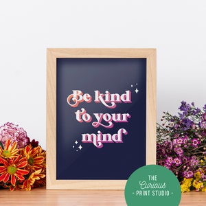 May include: A framed print with a navy blue background and white, pink, and orange text that reads "Be kind to your mind". The frame is light brown wood and there are small white stars around the text. The print is sitting on a wooden surface with flowers in front of it.