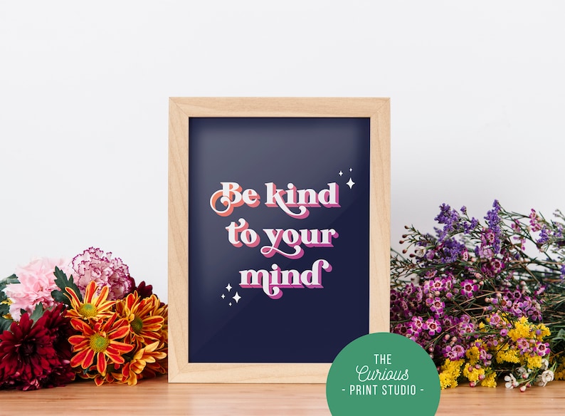 May include: A framed print with a navy blue background and white, pink, and orange text that reads "Be kind to your mind". The frame is light brown wood and there are small white stars around the text. The print is sitting on a wooden surface with flowers in front of it.