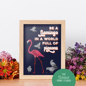Puede incluir: Una impresión enmarcada con un fondo azul y el texto "BE A flamingo IN A WORLD FULL OF pigeons". La impresión presenta un flamenco rosa de pie sobre una pierna con una bandada de palomas grises a su alrededor.