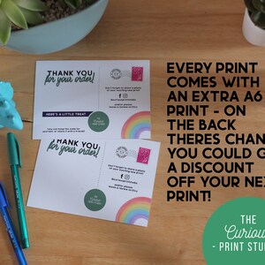 Puede incluir: Dos postales blancas con el texto "&iexcl;Gracias por tu pedido!" y "Aqu&iacute; hay un peque&ntilde;o obsequio" est&aacute;n sobre una superficie de madera. Las postales tienen un dise&ntilde;o de arco&iacute;ris y el texto "The Curious Print Studio". Tambi&eacute;n se ven dos bol&iacute;grafos y peque&ntilde;as plantas.