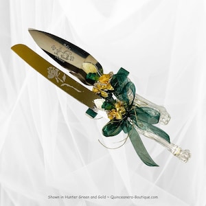 May include: A silver cake knife and server set with a green and gold ribbon and flower embellishment. The knife is engraved with "Mis 15 Años".
