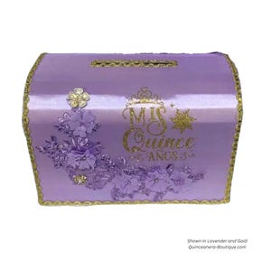 May include: A lavender satin keepsake box with gold trim and embellishments. The box has a slot on the top for money and features the text "Mis Quince Anos" in gold glitter.