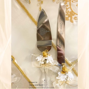 May include: Ivory and gold cake server and knife set. The stainless steel server has a decorative design and the knife is engraved with text. Both have floral and ribbon accents. The text on the image says "Shown in Ivory and Gold ~ Quinceanera-Boutique.com".