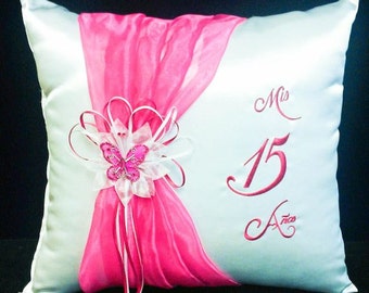 May include: White satin pillow with a pink sheer overlay and a pink butterfly embellishment. The pillow is embroidered with the text "Mis 15 Años".