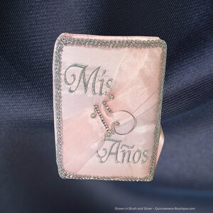 May include: A pink satin guest book with silver trim and a silver embroidered "Mis Quince Años" design. The book has a rhinestone embellishment in the shape of the number 15.