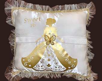May include: White satin pillow with gold accents and a decorative design. The pillow features a gold silhouette of a woman in a gown, a butterfly, and the text "Sweet 16". The edges are trimmed with gold and white lace.