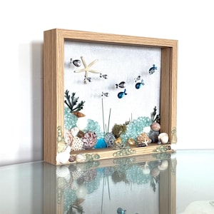 May include: A framed shadow box featuring a miniature ocean scene. The artwork includes a starfish, small metal fish, seashells, and sea glass. The frame is made of light-colored wood, and the scene is set against a white background.