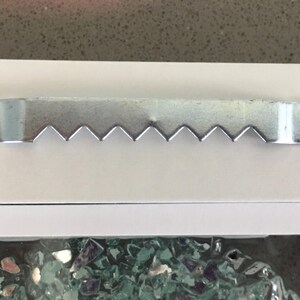 May include: A silver-colored metal sawtooth picture hanger attached to a white surface with two small nails. The hanger features a series of triangular teeth along the bottom edge, designed for mounting frames or wall decor securely.