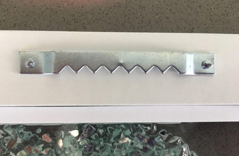 May include: A silver-colored metal sawtooth picture hanger attached to a white surface with two small nails. The hanger features a series of triangular teeth along the bottom edge, designed for mounting frames or wall decor securely.
