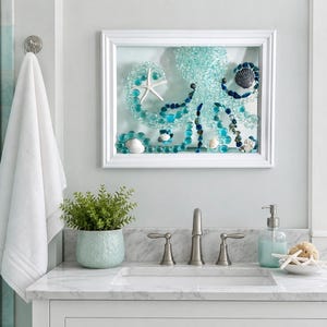 May include: A framed wall art piece featuring a mosaic octopus crafted from various shades of blue and clear glass beads, accented with a white starfish and dark shells, all encased within a simple white rectangular frame.