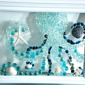 May include: A white framed wall art piece featuring an octopus mosaic crafted from light blue glass pebbles, dark blue stones, and various white and grey seashells. A white starfish is positioned on one of the octopus tentacles.