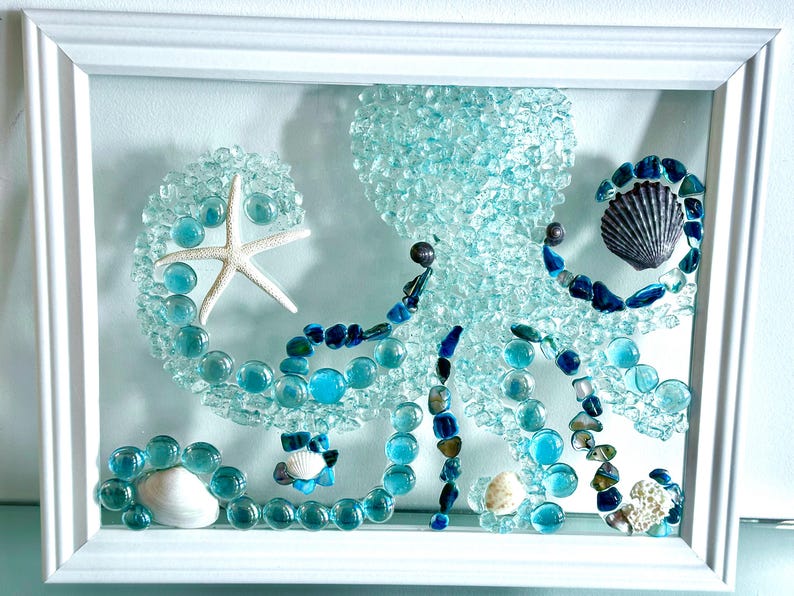 May include: A white framed wall art piece featuring an octopus mosaic crafted from light blue glass pebbles, dark blue stones, and various white and grey seashells. A white starfish is positioned on one of the octopus tentacles.