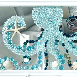 May include: A framed wall art piece featuring an octopus mosaic crafted from light blue glass shards, smooth turquoise glass pebbles, and dark blue stones. The design includes a white starfish, various seashells, and a white rectangular frame.