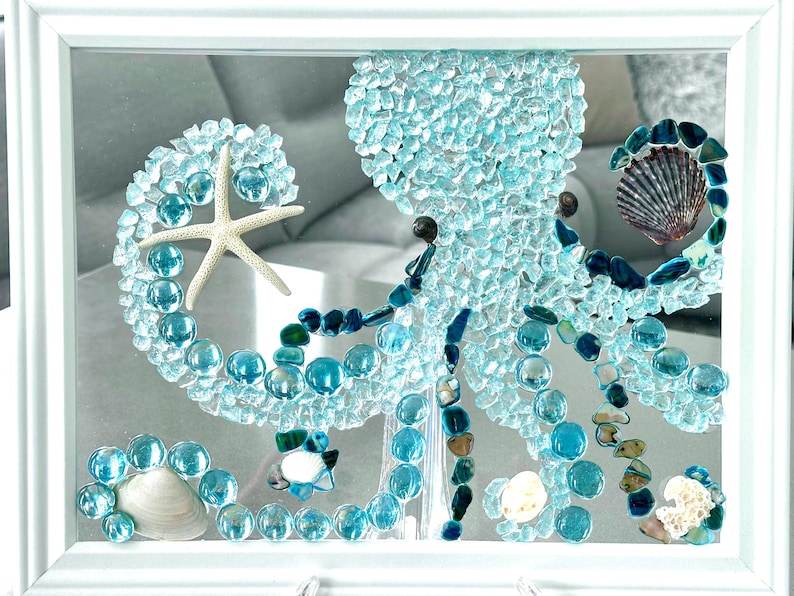 May include: A framed wall art piece featuring an octopus mosaic crafted from light blue glass shards, smooth turquoise glass pebbles, and dark blue stones. The design includes a white starfish, various seashells, and a white rectangular frame.