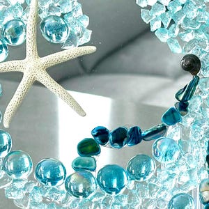 May include: A decorative piece featuring a white starfish surrounded by light blue glass pebbles and crushed turquoise glass fragments. The arrangement includes dark blue iridescent shell pieces and a small dark spiral seashell on a clear surface.