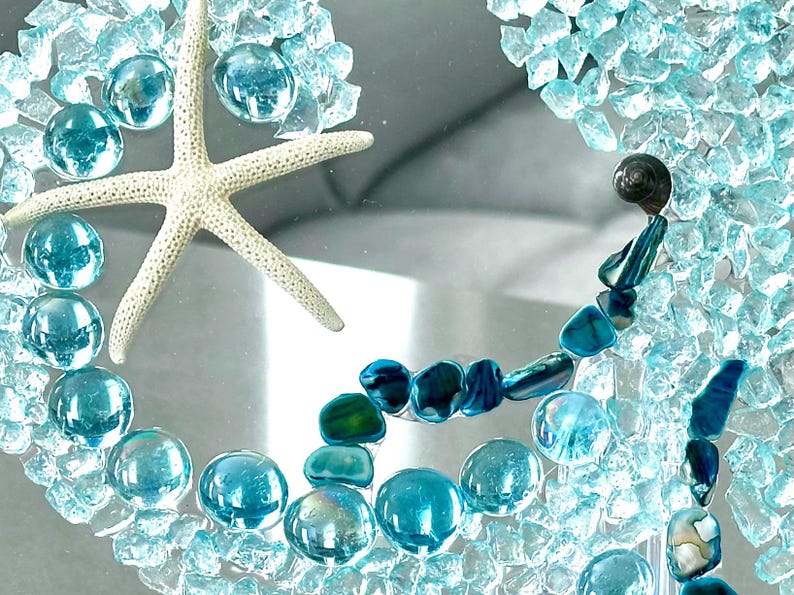 May include: A decorative piece featuring a white starfish surrounded by light blue glass pebbles and crushed turquoise glass fragments. The arrangement includes dark blue iridescent shell pieces and a small dark spiral seashell on a clear surface.