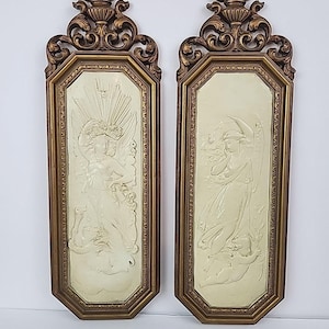 May include: A pair of vintage wall plaques with gold-coloured frames. Each plaque has a cream-coloured relief of a classical figure. The frames have a decorative top with a floral design. A classic home decor item.