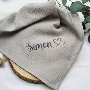 Könnte beinhalten: Eine hellgraue Babydecke mit dem Namen "Simon" in Schreibschrift gestickt, begleitet von einem Herzsymbol. Die Decke besteht aus einem weichen, strukturierten Stoff. Ein Schnuller und eine Holzscheibe sind ebenfalls zu sehen.