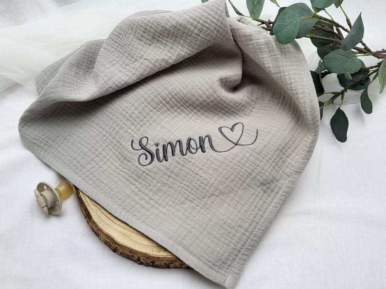 May include: A light gray muslin cloth featuring the embroidered name Simon in a dark gray cursive script followed by a heart outline. The fabric has a crinkled texture and is accompanied by a beige pacifier and a small wooden slice.