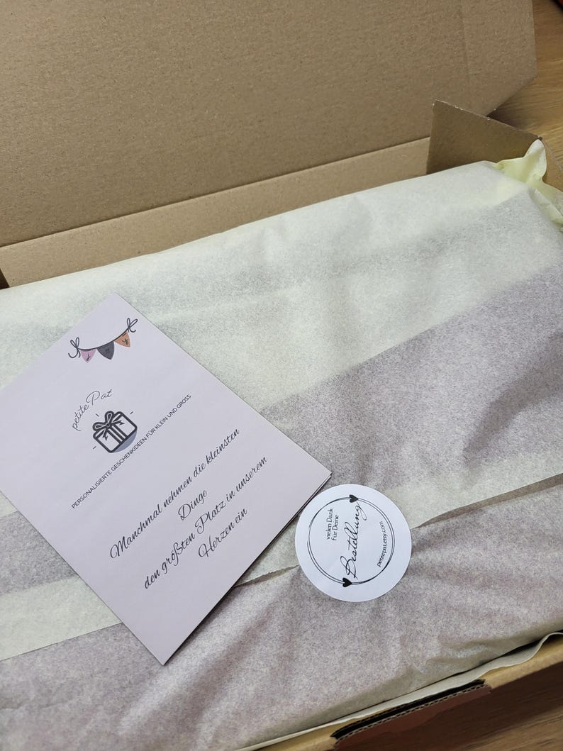 May include: A white box with a white tissue paper lining. The box contains a small card with the text "petite Pat PERSONALISERTE GESCHENKIDEEN F&Uuml;R KLEIN UND GROSS Manchmal nehmen die kleinsten Dinge den gr&ouml;&szlig;ten Platz in unserem Herzen ein" and a round sticker with the text "vielen Dank f&uuml;r Deine Bestellung".
