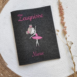 Könnte beinhalten: Ein grauer Filzordner mit dem Wort "Zeugnisse" in rosa gestickt. In der Mitte des Ordners ist ein rosa Ballerinamädchen gestickt. Der Name "Marie" ist unten auf dem Ordner in rosa gestickt.