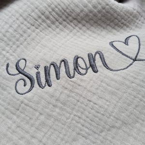 May include: A light gray muslin fabric featuring a textured, crinkled surface. The fabric is embroidered with the name Simon in a dark gray cursive script, followed by a simple, hand-drawn style heart outline in the same dark gray thread.