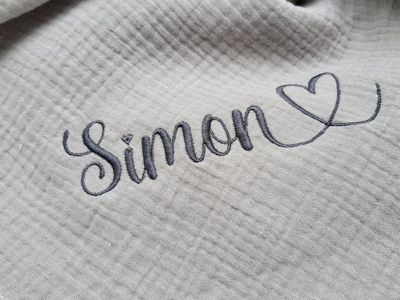 May include: A light gray muslin fabric featuring a textured, crinkled surface. The fabric is embroidered with the name Simon in a dark gray cursive script, followed by a simple, hand-drawn style heart outline in the same dark gray thread.