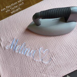 May include: A pink fabric with the name "Melina" embroidered in white with a heart next to it. A gray iron with a black handle is on top of the fabric. The text "zum Abschluss werden alle T&uuml;cher noch geb&uuml;gelt." is visible in the top left corner of the image.