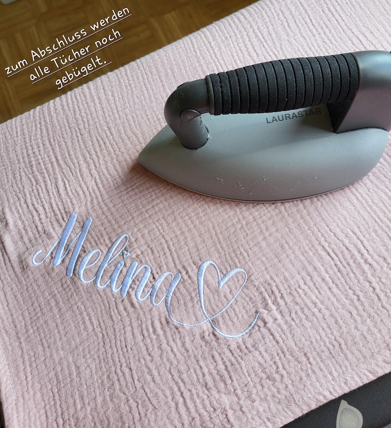 May include: A pink fabric with the name "Melina" embroidered in white with a heart next to it. A gray iron with a black handle is on top of the fabric. The text "zum Abschluss werden alle T&uuml;cher noch geb&uuml;gelt." is visible in the top left corner of the image.