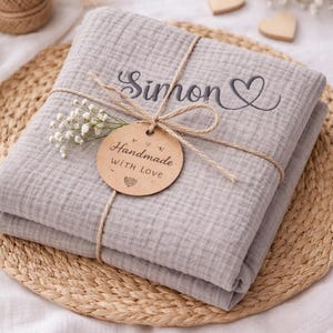 May include: A folded light gray muslin blanket featuring the embroidered name Simon and a heart symbol. The blanket is tied with twine and includes a round cardboard tag printed with the text Handmade WITH LOVE and a small heart icon.