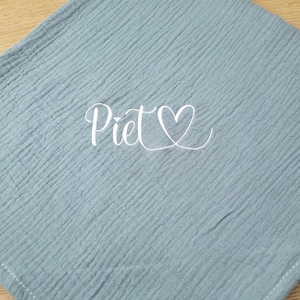 May include: A square, dusty blue muslin cloth featuring a crinkled texture and white stitched edges. The center of the fabric displays the name Piet in a white cursive script font, accompanied by a small white heart outline symbol.