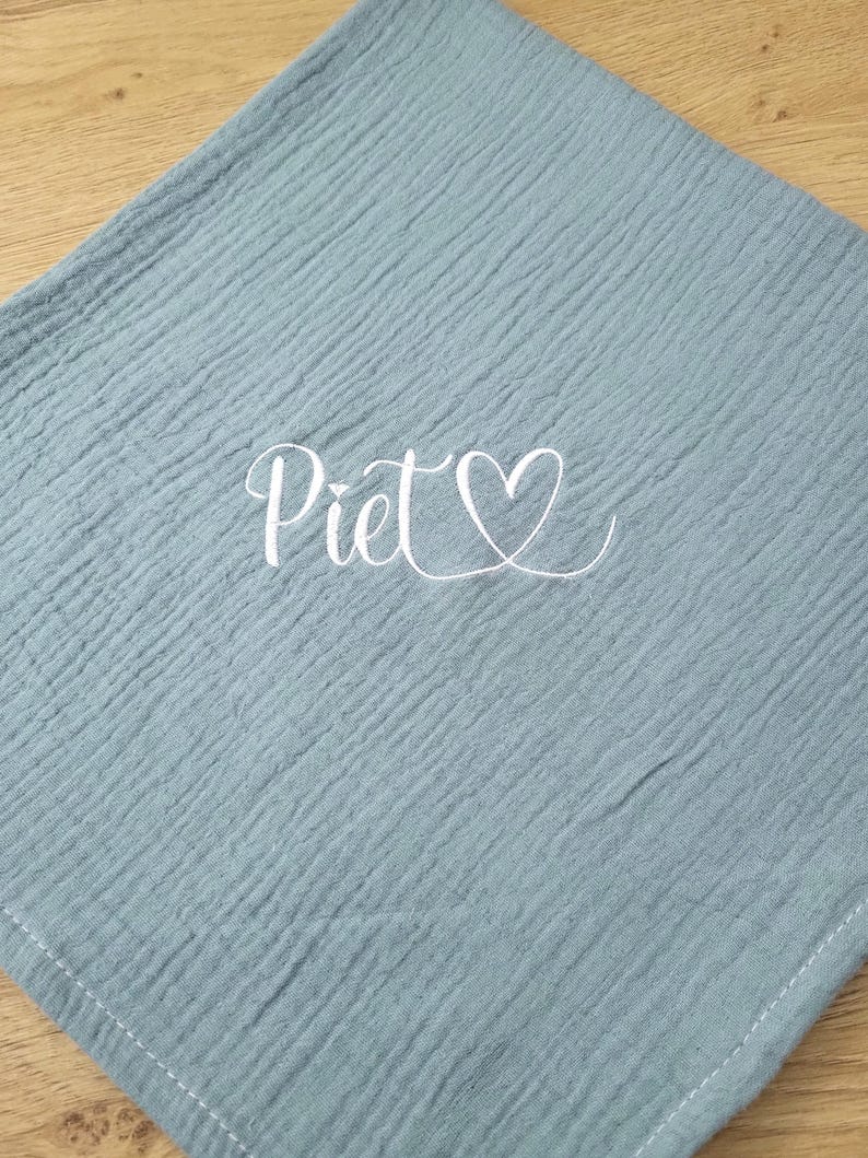 May include: A square, dusty blue muslin cloth featuring a crinkled texture and white stitched edges. The center of the fabric displays the name Piet in a white cursive script font, accompanied by a small white heart outline symbol.